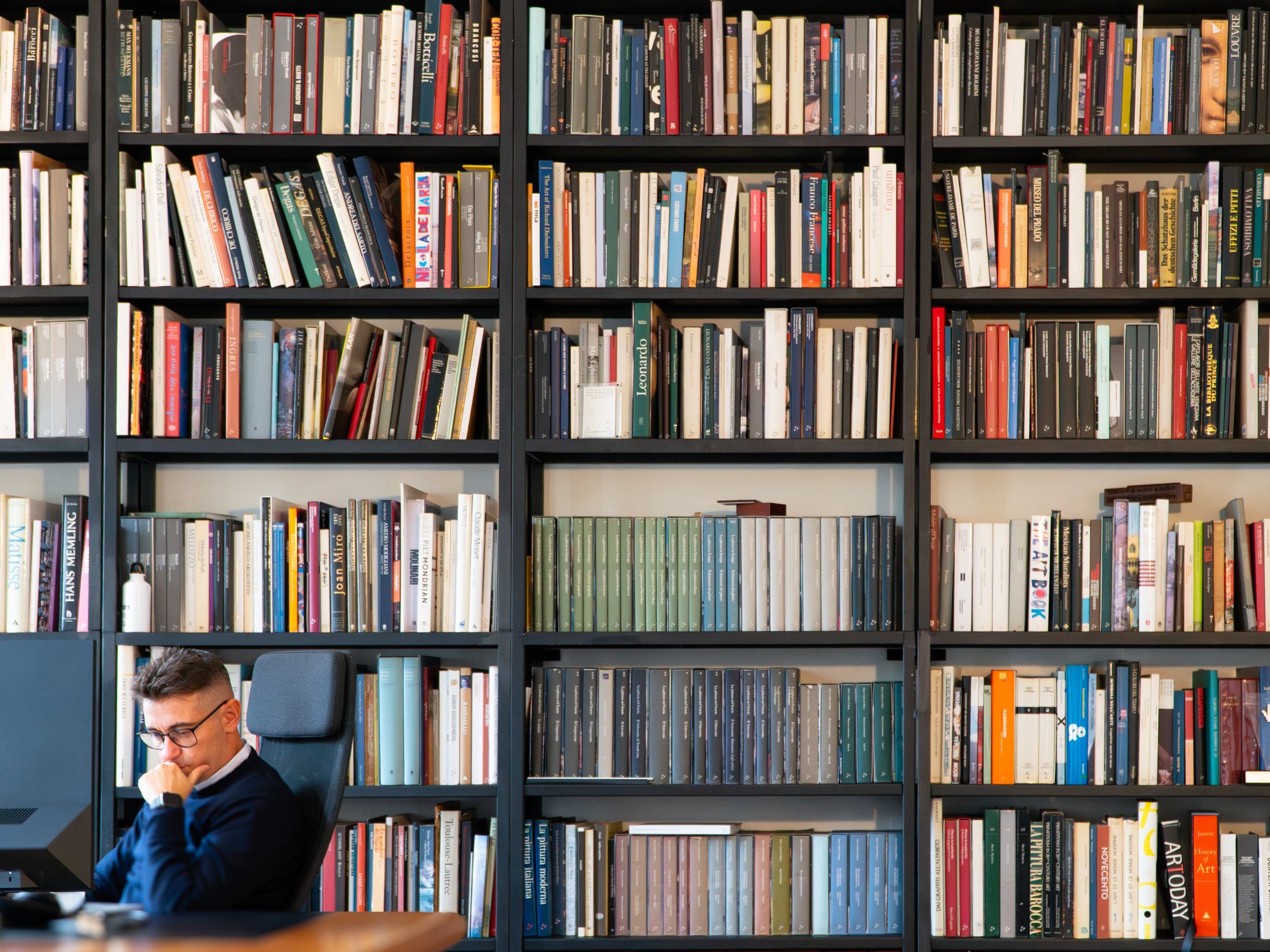 Uomo con occhiali seduto davanti a una libreria piena di libri.
