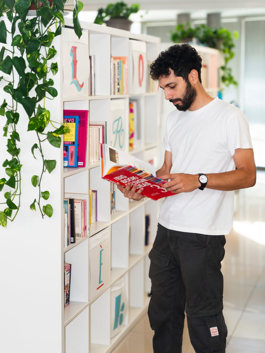 Uomo con barba e maglietta bianca legge un libro rosso in una libreria.