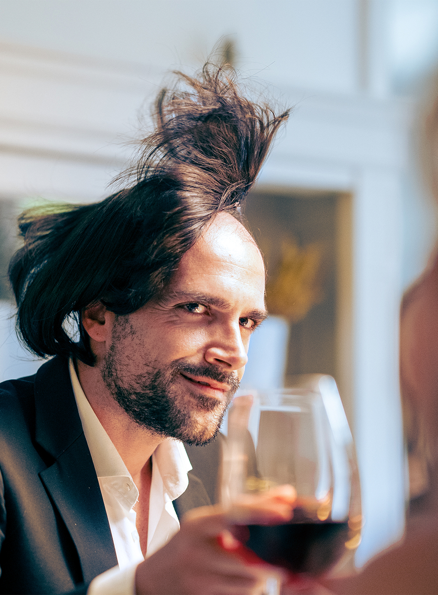 Primo piano di un uomo con barba che indossa un abito e tiene un bicchiere di vino rosso.