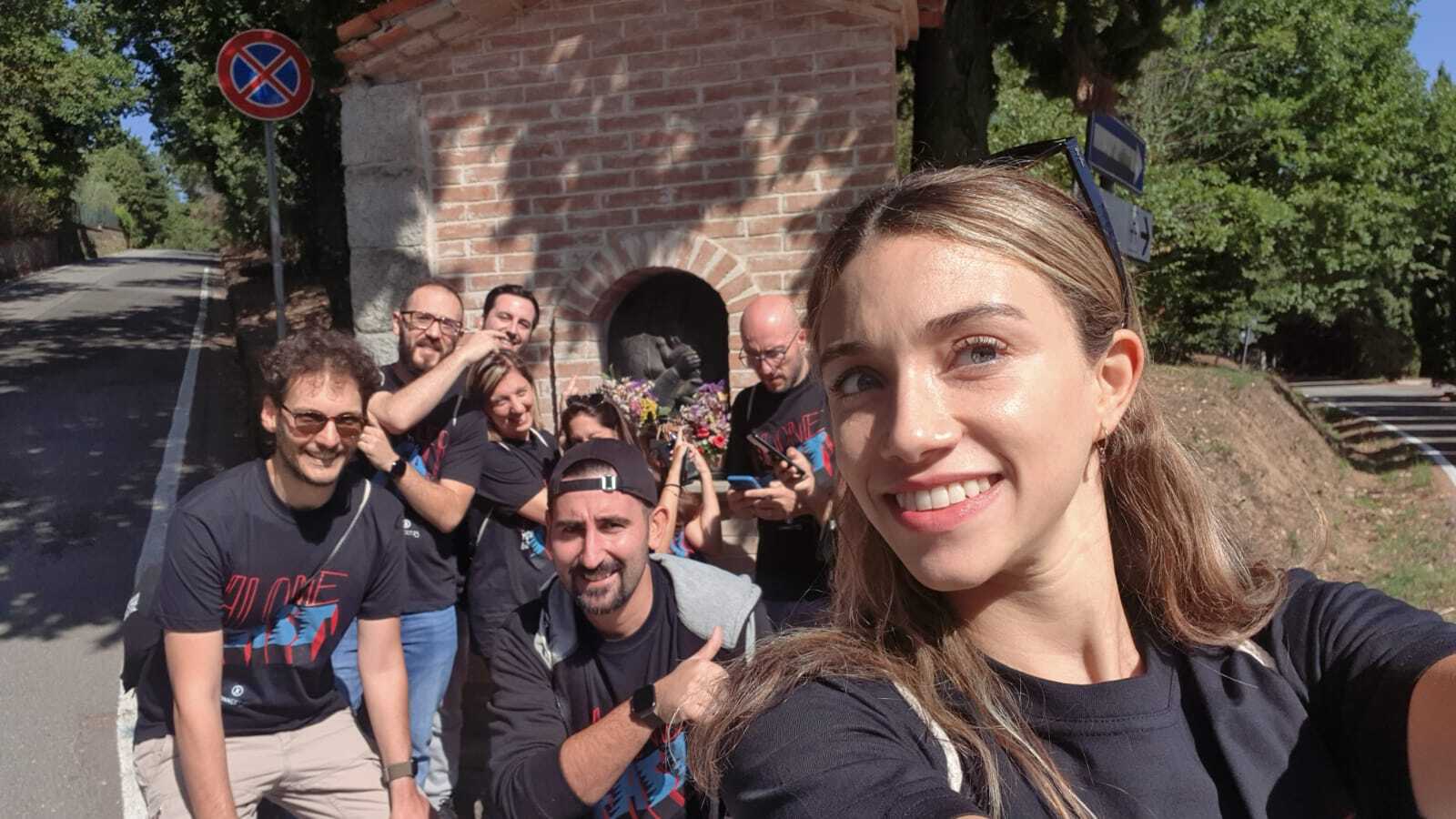 Selfie di gruppo di persone con magliette ALONE FIT vicino a una piccola edicola votiva.