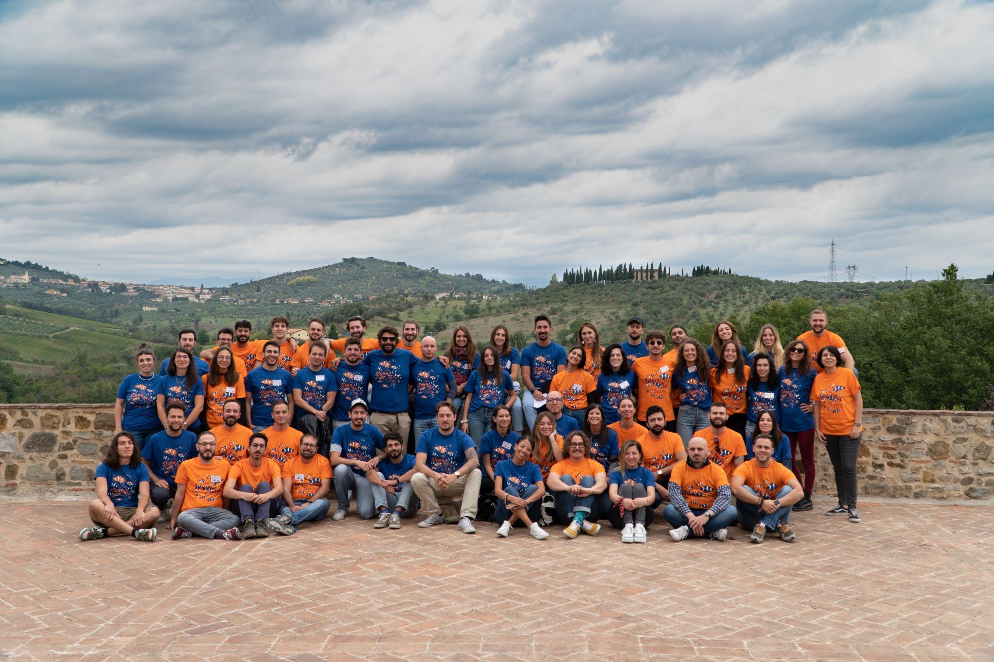 Folla di persone posa per una foto all'aperto, indossano sia magliette blu che arancioni.